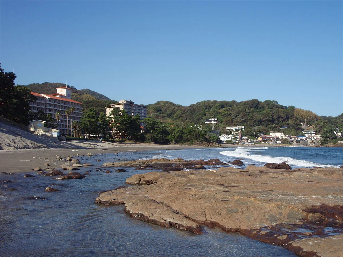 Rock shelves and water at the edge of Imaihama, with sand and wooded slopes behind, photographed in October 2011